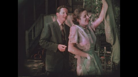 1950s: woman outside clicks film slate, woman hangs clothes on clothesline, takes clothespin from apron pocket while man in suit talks, woman laughs, talks, reaches for clothes