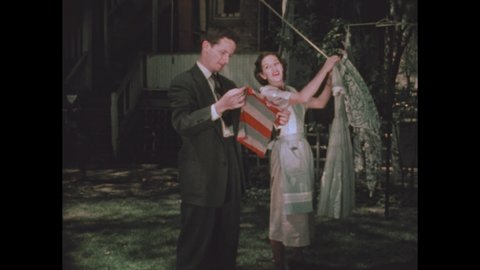 1950s: man in suit kneels by laundry basket, gives dress to woman near clothesline, they talk, she takes clothespin from apron pocket, hangs dress, man gets shirt from basket, hands it to woman