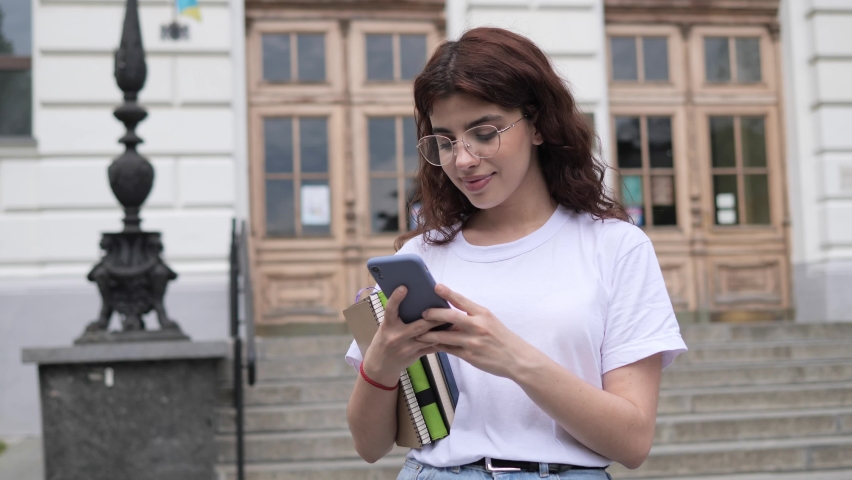 Positive female curly hair student in white t-shirt with books adjusts glasses on the background of college outdoor. Beautiful girl looking to camera. Happy face of young woman with healthy smile.