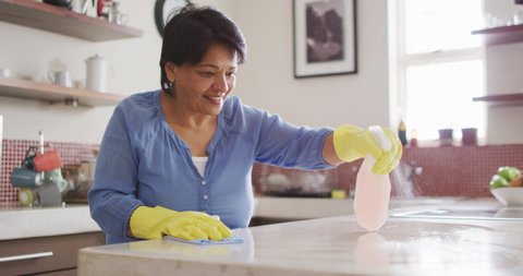 Smiling senior biracial woman wearing gloves and cleaning table in kitchen alone. healthy and active retirement at home.