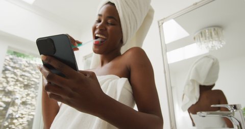 Video of happy african american woman in robe brushing teeth and using smartphone. beauty treatment and skin care routine concept.