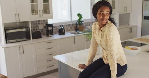 Video of happy african american woman sitting on countertop in kitchen. leisure, relax and spending free time at home.