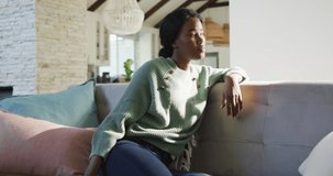 Video of thoughtful african american woman sitting on sofa and looking outside window. lifestyle and spending free time at home.