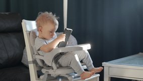 Cute kid sitting in booster seat fixed on top of dining chair, baby playing with mobile phone. High quality 4k footage