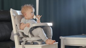 Cute kid with food nibbler baby fruit pacifier sitting in booster seat fixed on top of dining chair watching tv. High quality 4k footage