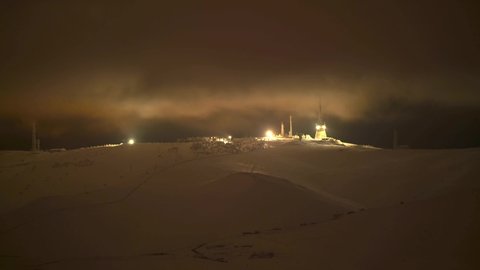 8K 7680x4320.Night lights of antennas on top of snowy mountain in fog and clouds.Light of transmitter antenna.Midnight mountain dark creepy background landscape nature.Hill ridge foggy peak summit top