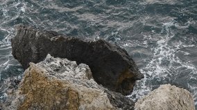 High angle view of dark sea waves crashing with splashes on large limestone rock on sea shore. Selective focus. Slow motion video. Bad weather forecast theme.
