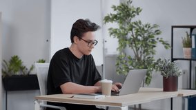 Modern education concept. Young guy student in eyeglasses typing on laptop and drinking takeout coffee, studying at home, slow motion, free space