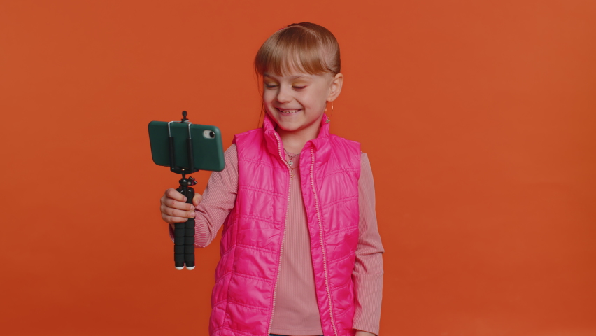 Girl child toddler blogger taking selfie on mobile phone selfie stick, communicating video call online with subscribers. Young little children kid teen isolated alone on orange studio wall background