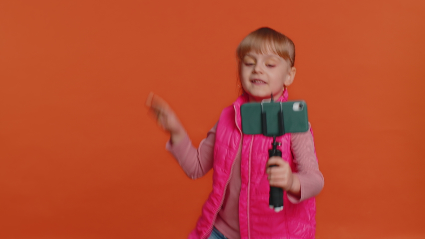 Girl child toddler blogger taking selfie on mobile phone selfie stick, communicating video call online with subscribers. Young little children kid teen isolated alone on orange studio wall background