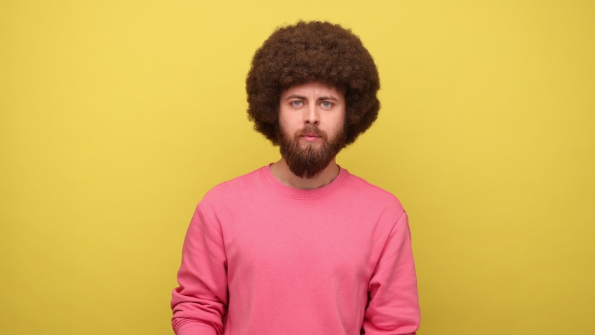 Bearded hipster man with Afro hairstyle pointing at mobile phone with blank screen, surprised facial expression, wearing pink sweatshirt. Indoor studio shot isolated on yellow background. Royalty-Free Stock Footage #1090254829