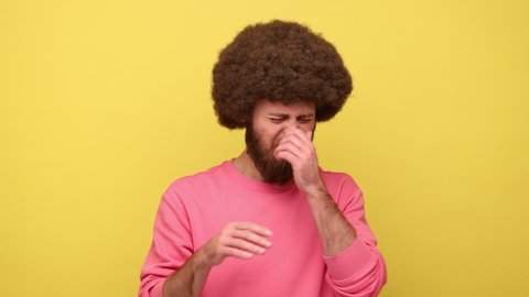 Man with Afro hairstyle holding breath with fingers on nose, grimacing in disgust, expressing repulsion to stink, fart gases, wearing pink sweatshirt. Indoor studio shot isolated on yellow background.