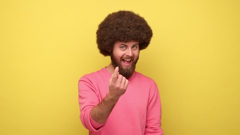 Bearded man with Afro hairstyle making beckoning gesture, inviting for date and looking alluring, flirting to camera, wearing pink sweatshirt. Indoor studio shot isolated on yellow background.