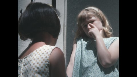1960s: Blonde girl pulls her finger out of her eye and talks to brunette girl with a bob hairstyle.