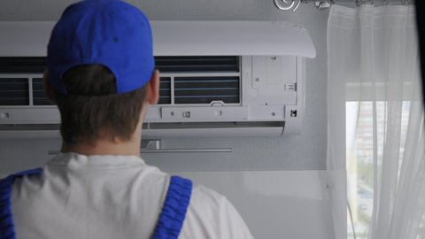 A professional repairman of air conditioning equipment replaces the filter elements. A man in uniform and gloves takes out the grilles from the air conditioner for cleaning and prevention