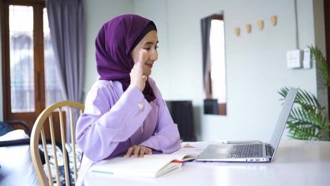 A Muslim woman wearing a hijab sits at a table in a cafe working with a laptop and a notepad.