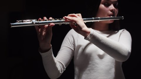 Young female student plays studio flute with dark background, front view. Woman performs music on flute in studio. Concept of art