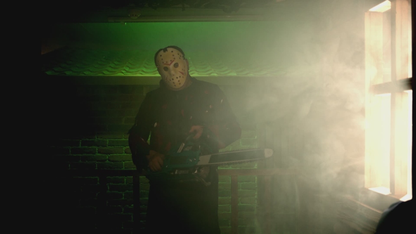 Silhouette of man holding chainsaw and walking inside dark smoked place . Portrait of scary man with vintage hockey mask in horror halloween scene . Friday 13th theme . Looking at camera . Close up | Shutterstock HD Video #1090260627