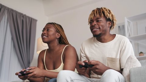 Home video game. Friends leisure. Fun weekend. Virtual challenge. Excited guy bothering girl trying to win playing with gamepad on couch in modern living room interior.