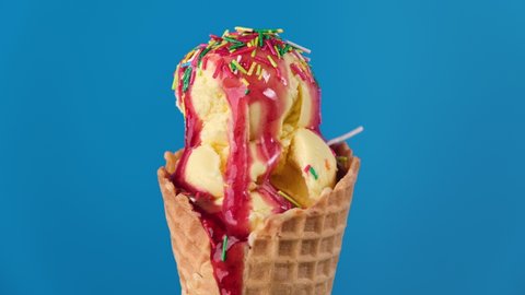 Timelapse of banana ice cream with strawberry topping and colorful sprinkles in waffle cone melting on blue background. Delicious yellow ice cream melting. Close-up of sweet dessert. Food concept