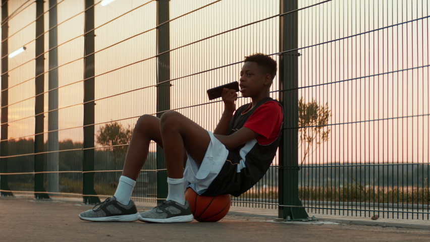 African American teenager sits on a ball and using his phone on basketball court. Shot with 2x anamorphic lens | Shutterstock HD Video #1090263305