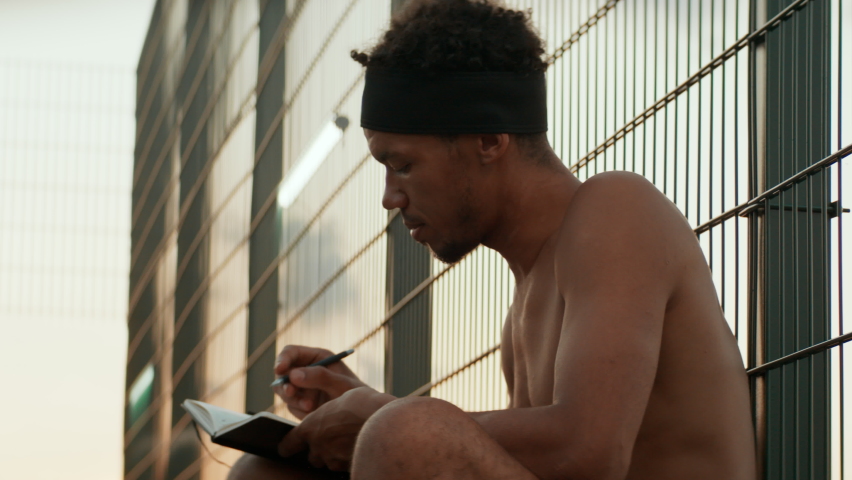 Young African American male writing lyrics for his song on a basketball court. Aspiring rap artist dreaming of becoming future star. Shot with 2x anamorphic lens | Shutterstock HD Video #1090263313