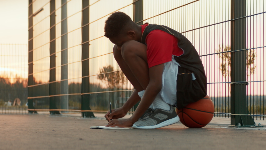Black African American teenager boy writing lyrics for his song on a basketball court. Aspiring rap artist dreaming of becoming future star. Shot with 2x anamorphic lens | Shutterstock HD Video #1090263321