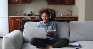 Confident gen z African female student learn study online on distance sit on sofa hold laptop communicate with teacher by video call using wireless headset take notes. Young woman get remote education