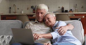 Friendly bonding older age husband wife spend time at cozy home use laptop computer read news at internet site make purchase online. Happy couple grandparents retirees cuddle on sofa look on pc screen