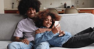 Laughing young female wife relax on sofa lying in warm embraces of beloved male husband hold cell show spouse funny photo video at social networks. Happy Black married couple rest indoors using phone