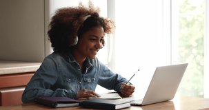 Busy African teen female high school pupil college student sit at table by laptop in headphones listen to video lesson at home write up information. Gen z lady prepare essay homework using modern tech