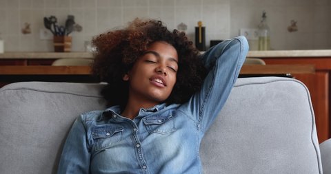 Calm millennial Afro American woman enjoy peaceful rest on sofa nap dream with closed eyes then look aside with smile imagine good future. Happy teenage female relax feel peace of mind relieve stress