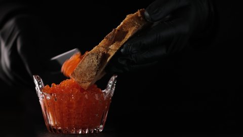 Process of making sandwiches with salmon caviar salted roe. Home cooking. Spreads red caviar on bread with spoon. Chef smears salmon caviar on toast, close-up black background slow motion