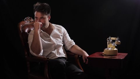 a man on black in a white shirt sits in a chair, in front of a retro telephone and cannot decide to pick up the phone