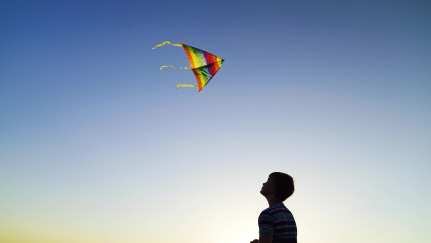 Boy plays kite with toy. Flying kite in blue sky, silhouette of child. Childhood, dream of flying, freedom travel. Happy boy playing with multi colored flying kite in nature in rays of summer sun.