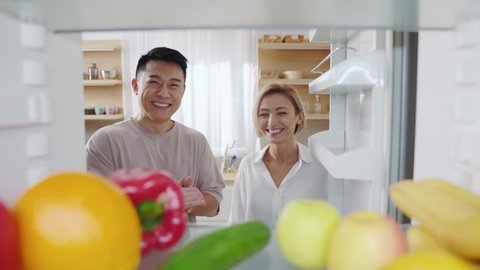 Footage of beautiful Caucasian girl with blonde hair opening door of refrigerator. Happy multiethnic couple in love looking inside, smiling. View from fridge, shelf with fruits and vegetables. Indoors