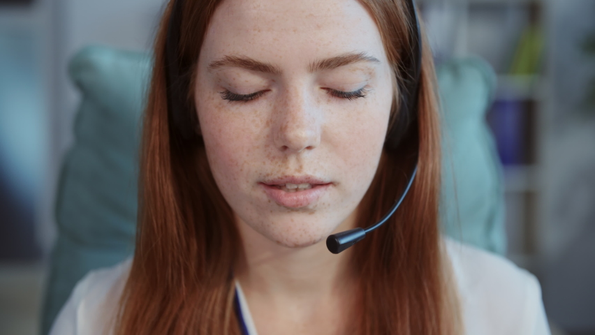 Close-up footage of cheerful young woman with ginger hair wearing headset looking art camera. Wonderful smiling girl in formal clothes sitting at workplace, looking at camera in call center. Indoors Royalty-Free Stock Footage #1090271195