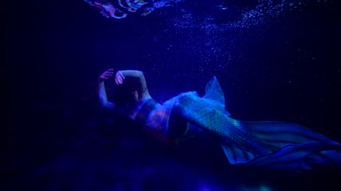 a mermaid is sitting on the bottom in blue water. the streaks of red light are sliding. the general plan