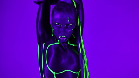 Portrait of confident woman with fluorescent makeup in bright costume dancing robotic dance looking at camera. Live camera zoom out as slim flexible dancer performing in ultraviolet light posing
