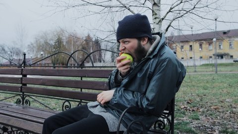 Hungry bearded homeless man sits on a bench and eats a green apple in a city park. Below poverty line. Male tramp in dirty clothes with cap and hat. Beggar. Illegal immigrant. Drunkenness, alcoholism