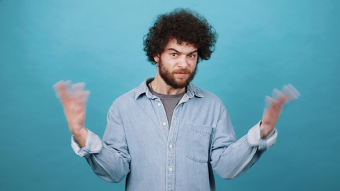 Young bearded guy in denim jacket shows hands gesture making person guess. Confident man with dark kinky hair stands on blue background
