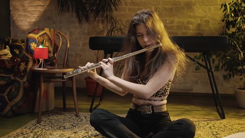 Zoom in portrait of young cute woman musician rehearsing music performance at home, playing flute, sitting on floor, slow motion