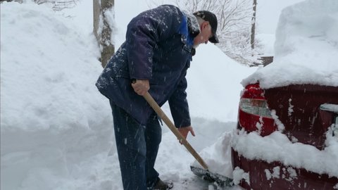 A man in a baseball cap, jacket, and jeans is clearing snow next to a red car. Clearing the area in winter after a snow storm. Shovel throws snow on a huge snowdrift. Snow falls from the sky