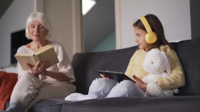 Positive girl with headphones looking at the tablet and talking with her grandmother reading book at home