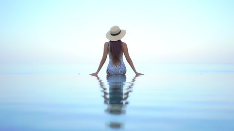 Back to the camera. A woman in a bathing suit and straw sun hats sits on the edge of an infinity pool. Title space