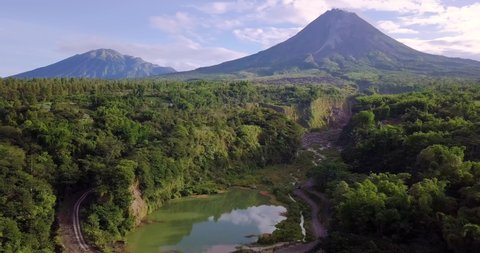 Aerial backwards flight showing natural lake surrounded by wilderness of Indonesia and giant Merapi Volcano in background during summer - Bego Pendem,Indonesia