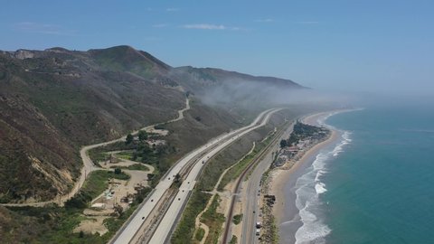wide aerial view of pacific coast highway route one as light traffic drives along the highway with mountains on one side and pacific ocean on the other on a sunny summer day