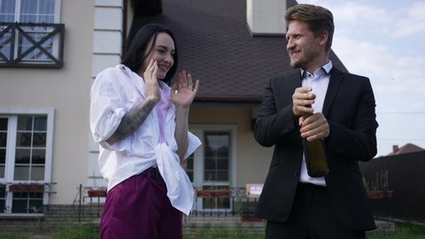 Excited rich couple opening champagne in front of new house outdoors. Positive happy wealthy Caucasian husband and wife celebrating real estate property purchase. Slow motion