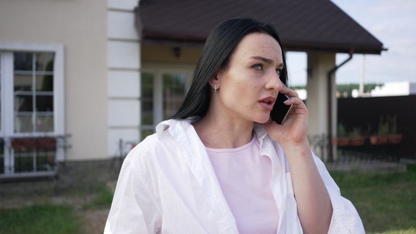 Worried troubled Caucasian businesswoman talking on phone standing on backyard outdoors. Portrait of attractive woman discussing business in slow motion on sunny morning. Lifestyle concept