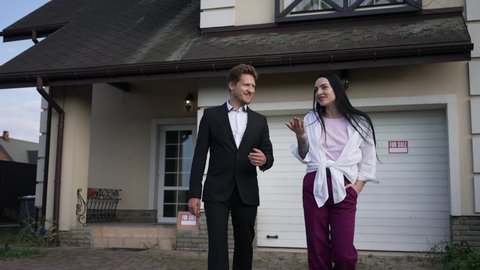 Confident wealthy couple walking in slow motion discussing purchase of new house for sale. Front view rich Caucasian man and woman strolling outdoors talking smiling. Lifestyle and relationship